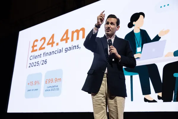 Liam Randall delivering a keynote on financial inclusion at Merchant Taylors' Hall — slide behind reads £24.4m client financial gains, 2025/26.