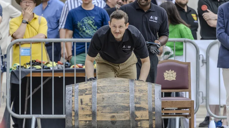 Liam Randall in a black polo shirt, bent forward rolling a large wooden cask along the ground, with a crowd watching from behind a barrier
