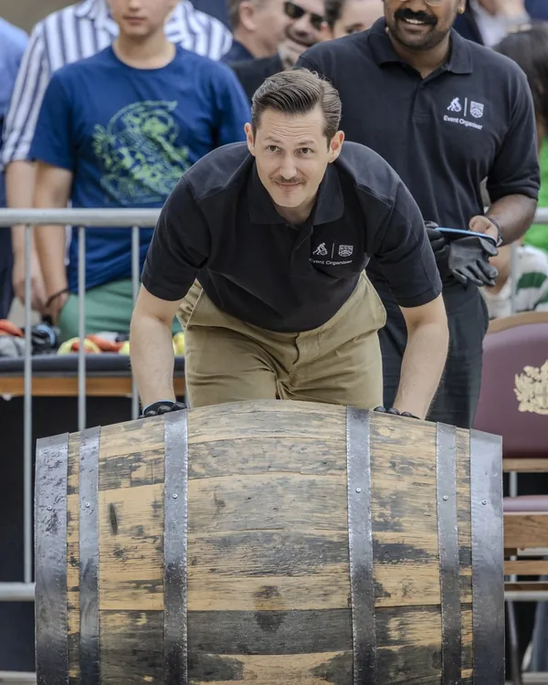 Liam Randall in a black polo shirt, bent forward rolling a large wooden cask along the ground, with a crowd watching from behind a barrier
