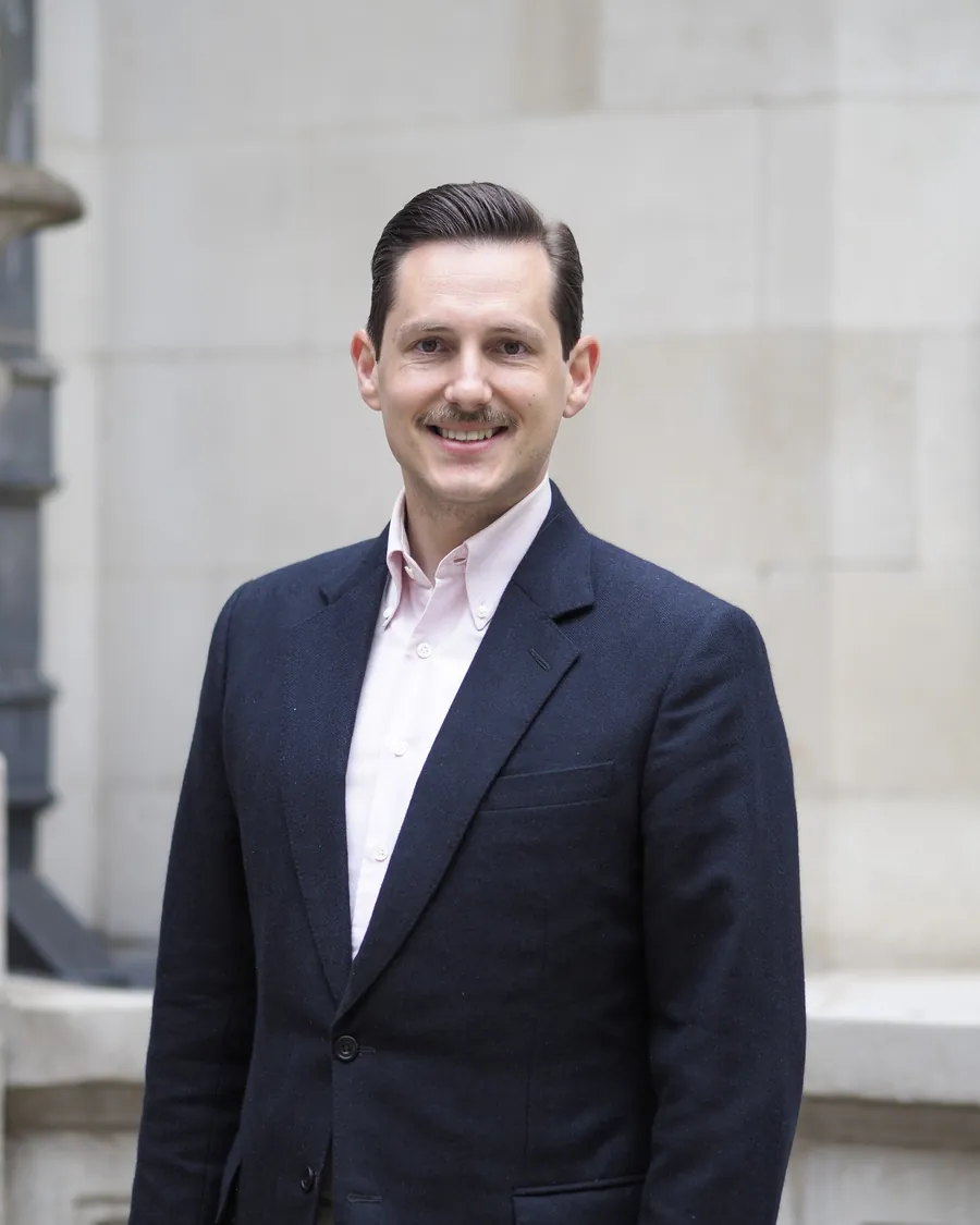 Liam Randall in a navy jacket and open-collared shirt, smiling, in front of a Portland stone wall in the City of London