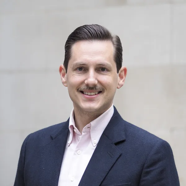 Liam Randall in a navy jacket and open-collared shirt, smiling, in front of a Portland stone wall in the City of London