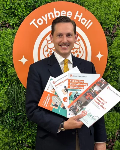 Liam Randall in a suit and tie, smiling, holding four Debt Free Advice impact reports in front of a Toynbee Hall sign